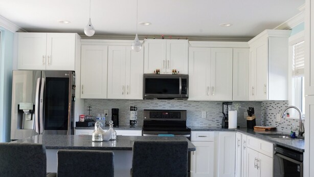 A modern kitchen with white cabinets, a stainless steel refrigerator, a microwave, and a gas stove. There are three bar stools and a large island with a sink. - Charming Home with Beautiful Backyard near Downtown  (Fort Myers)