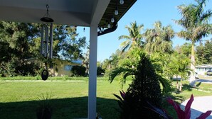 A covered patio area with hanging light fixtures, a potted plant, and a view of a well-maintained lawn and palm trees. - Charming Home with Beautiful Backyard near Downtown  (Fort Myers)