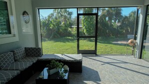 A spacious living area with a sectional sofa, a coffee table with plants, and a large sliding glass door leading to a grassy outdoor area. - Charming Home with Beautiful Backyard near Downtown  (Fort Myers)