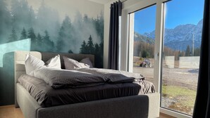 Ein Schlafzimmer mit einem großen Bett, Blick auf eine gebirgige Landschaft durch eine Schiebetür und einem Waldgemälde an der Wand.