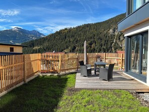 A wooden deck with outdoor furniture, a view of a mountain range, and a modern building with large windows. - Chalet apartment with panoramic mountain views at the foot of the Zugspitze  (Ehrwald)