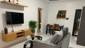 A modern living room with a grey sofa, a TV mounted on the wall, a wooden shelf, a dining table with chairs, and a potted plant. - The house of happiness (Abidjan)