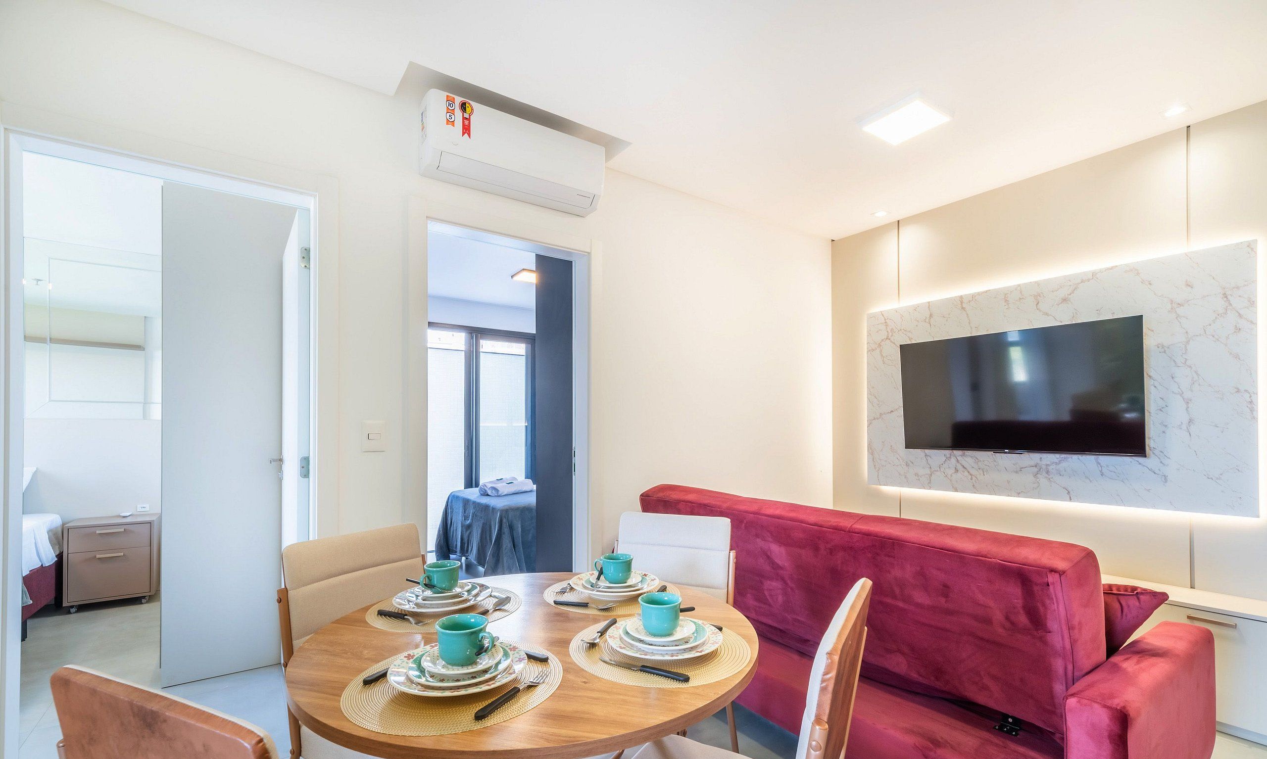 A dining area with a round table set for four, a red sofa, and a flat-screen TV mounted on the wall.