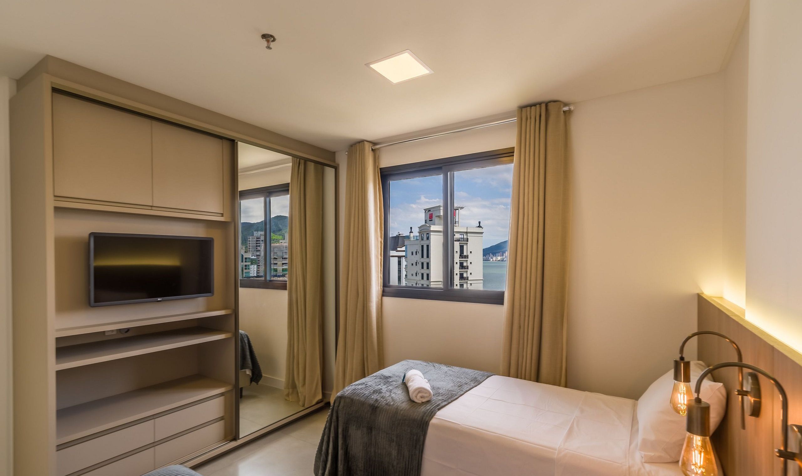 A hotel room with a bed, a TV, a wardrobe, and a window with a city view.