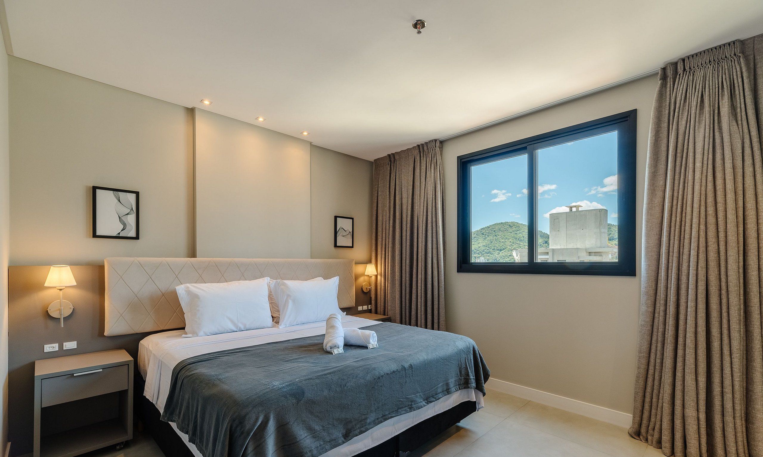Una habitación de hotel con cama, mesitas de noche, ventana con cortinas y cuadros en la pared.