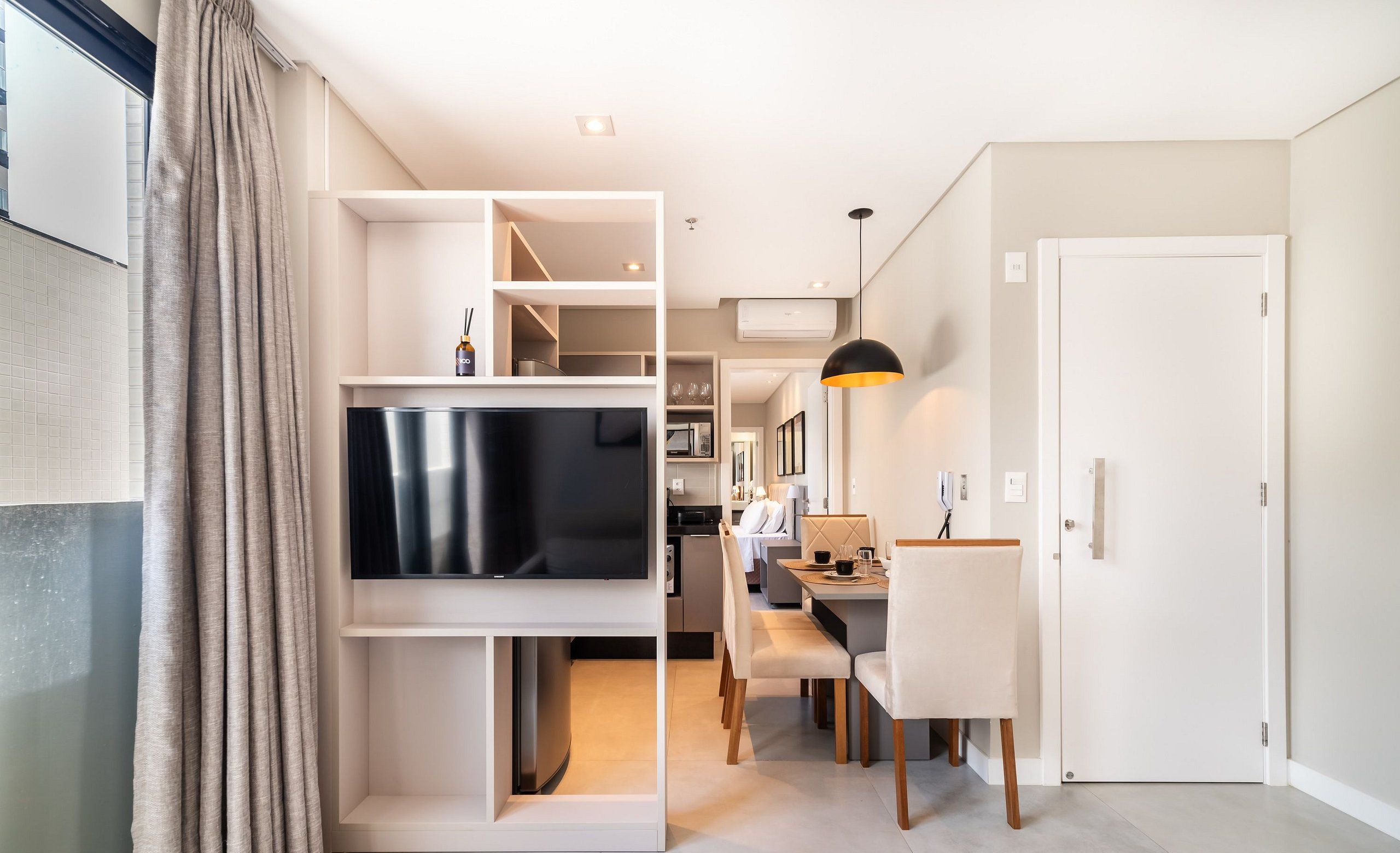 Una sala de estar moderna con un televisor de pantalla plana montado en una pared blanca, una zona de comedor con mesa y sillas, y una cocina al fondo.