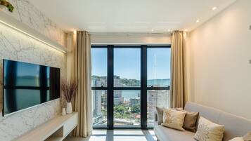 Una sala de estar moderna con un televisor grande, un sofá con cojines decorativos y vistas a un paisaje urbano a través de ventanas de piso a techo.