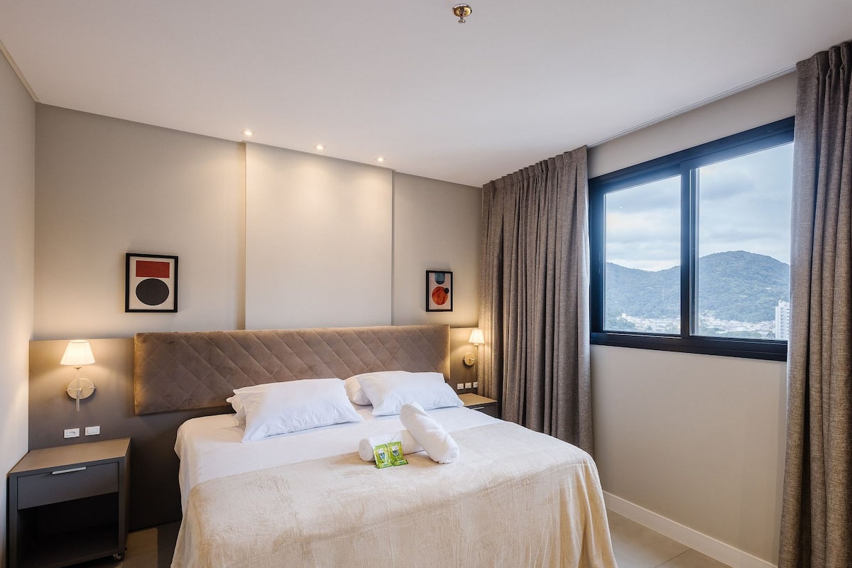 Une chambre d’hôtel avec un lit, des tables de chevet, des œuvres d’art murales, une fenêtre avec des rideaux et une vue sur une montagne.