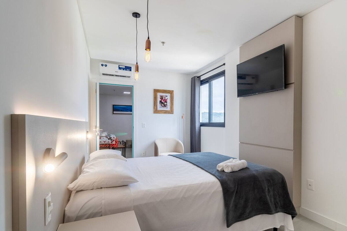 Une chambre d’hôtel moderne équipée d’un lit, d’une table de chevet, d’un téléviseur fixé au mur et d’une fenêtre donnant sur un paysage agréable.