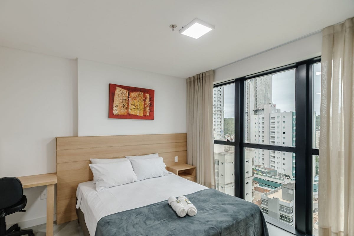 Une chambre d’hôtel équipée d’un lit, d’un bureau, d’une chaise, d’une fenêtre donnant sur la ville et d’un luminaire au plafond.