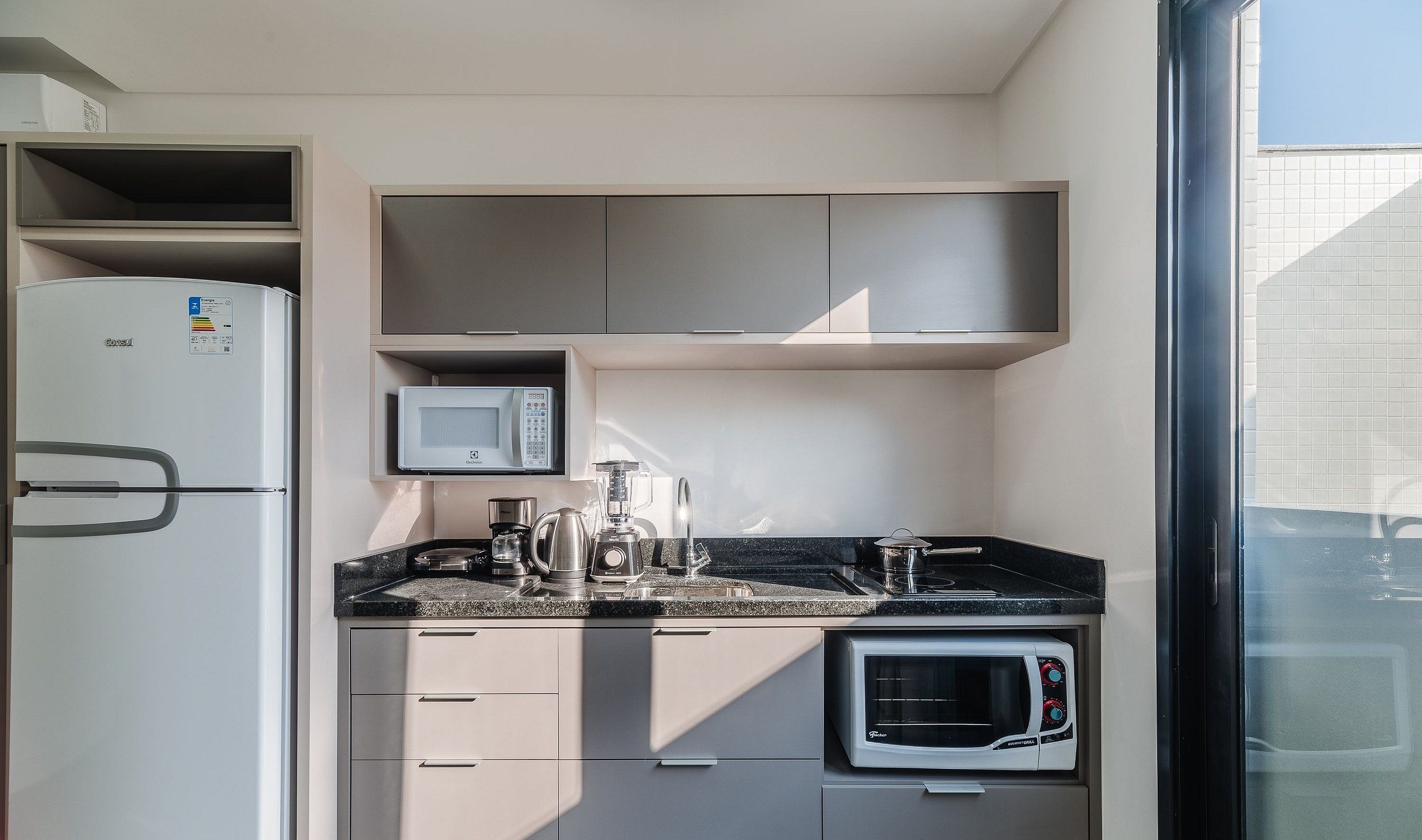 A compact kitchen with a refrigerator, microwave, kettle, blender, and a built-in oven.