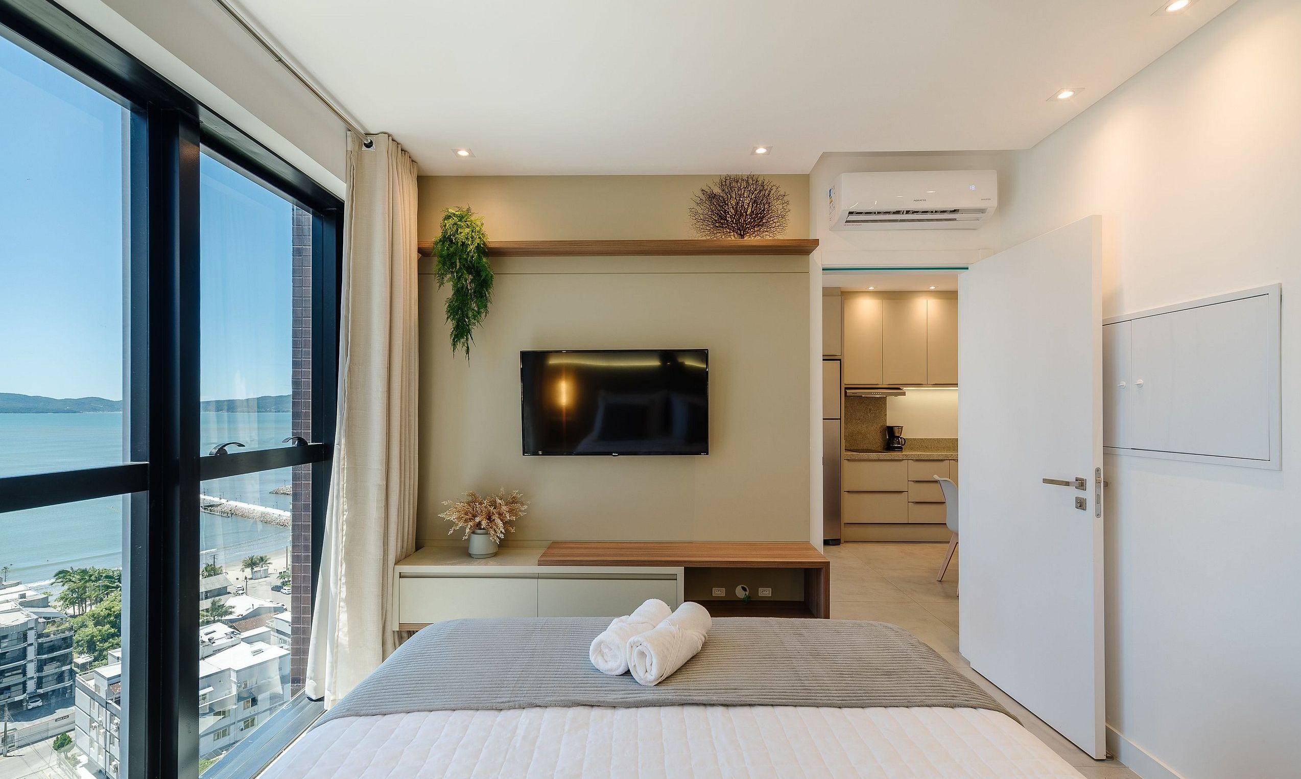 A modern bedroom with a large window, a flat-screen TV, a bedside table with a plant, and a view of the sea.