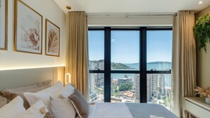 A bedroom with a large window, a bed with multiple pillows, and framed artwork on the wall. - MHF III Apts Variados a 280m da Praia em Itapema (Itapema)