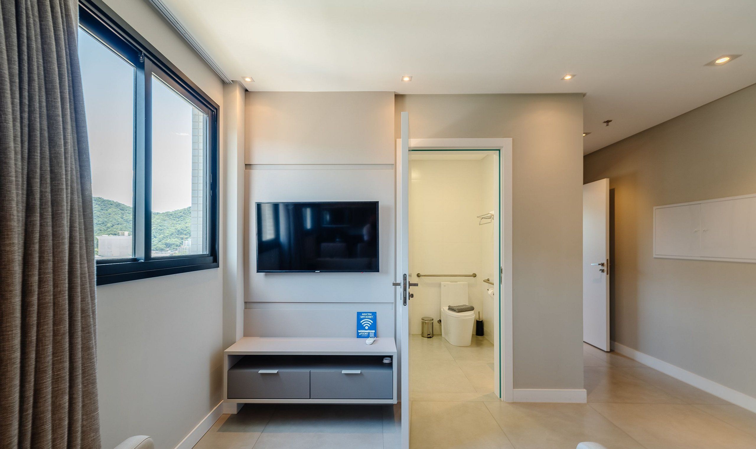 A modern hotel room with a large window, a flat-screen TV mounted on the wall, a small seating area with storage, and a door leading to a bathroom.