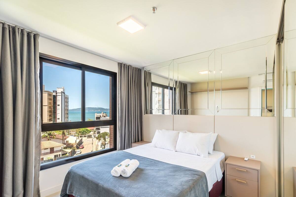 Une chambre d’hôtel avec un grand lit, une table de chevet, un miroir et une vue sur des immeubles et une étendue d’eau.