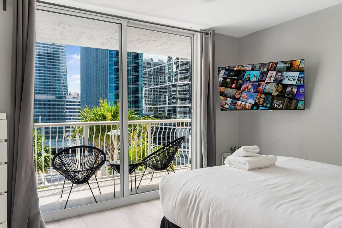 A modern hotel room with a large balcony, a bed with white linens, and a wall-mounted TV displaying movie posters.
