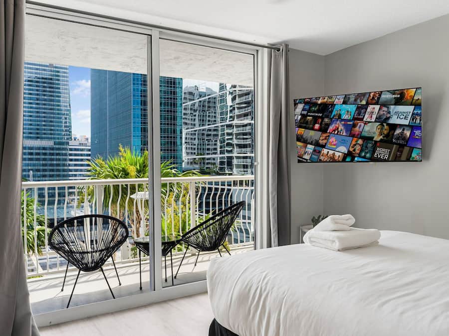 Quarto de hotel moderno com uma varanda grande, cama com roupa de cama branca e televisão fixada na parede exibindo cartazes de filmes.