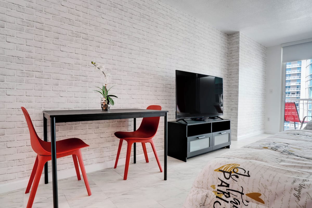 A modern room with a white brick wall, a black TV stand, a dining table with red chairs, and a bed with a patterned cover.