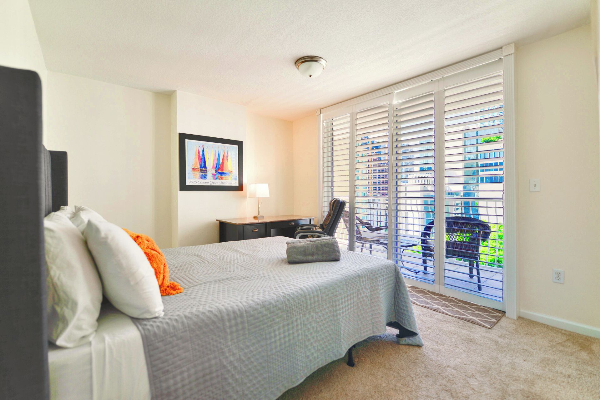 A bedroom with a bed, a desk, a chair, a framed picture on the wall, and a view of the outside through large windows.