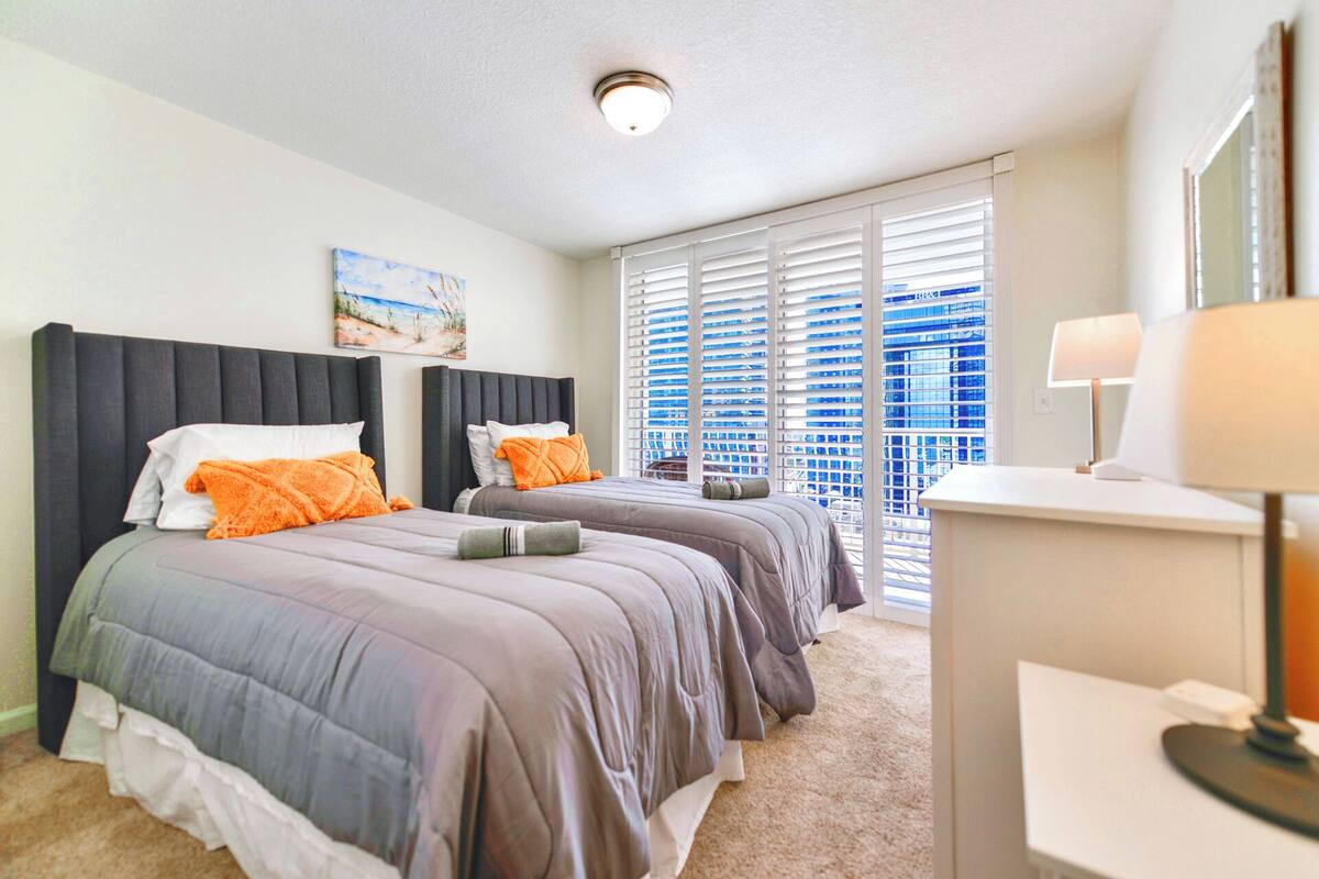 A hotel room with two beds, a painting on the wall, a window with blinds, and a desk with a lamp.