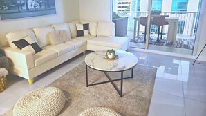 Una sala de estar moderna con un sofá seccional blanco, una mesa de centro redonda y un ventanal con vistas a un edificio.