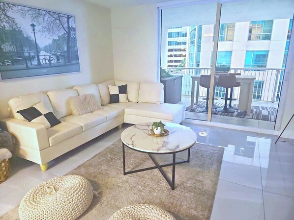 A modern living room with a white sectional sofa, a round coffee table, and a large window offering a view of a building. - 1200 Brickell - Miami stay - We Host (Miami)