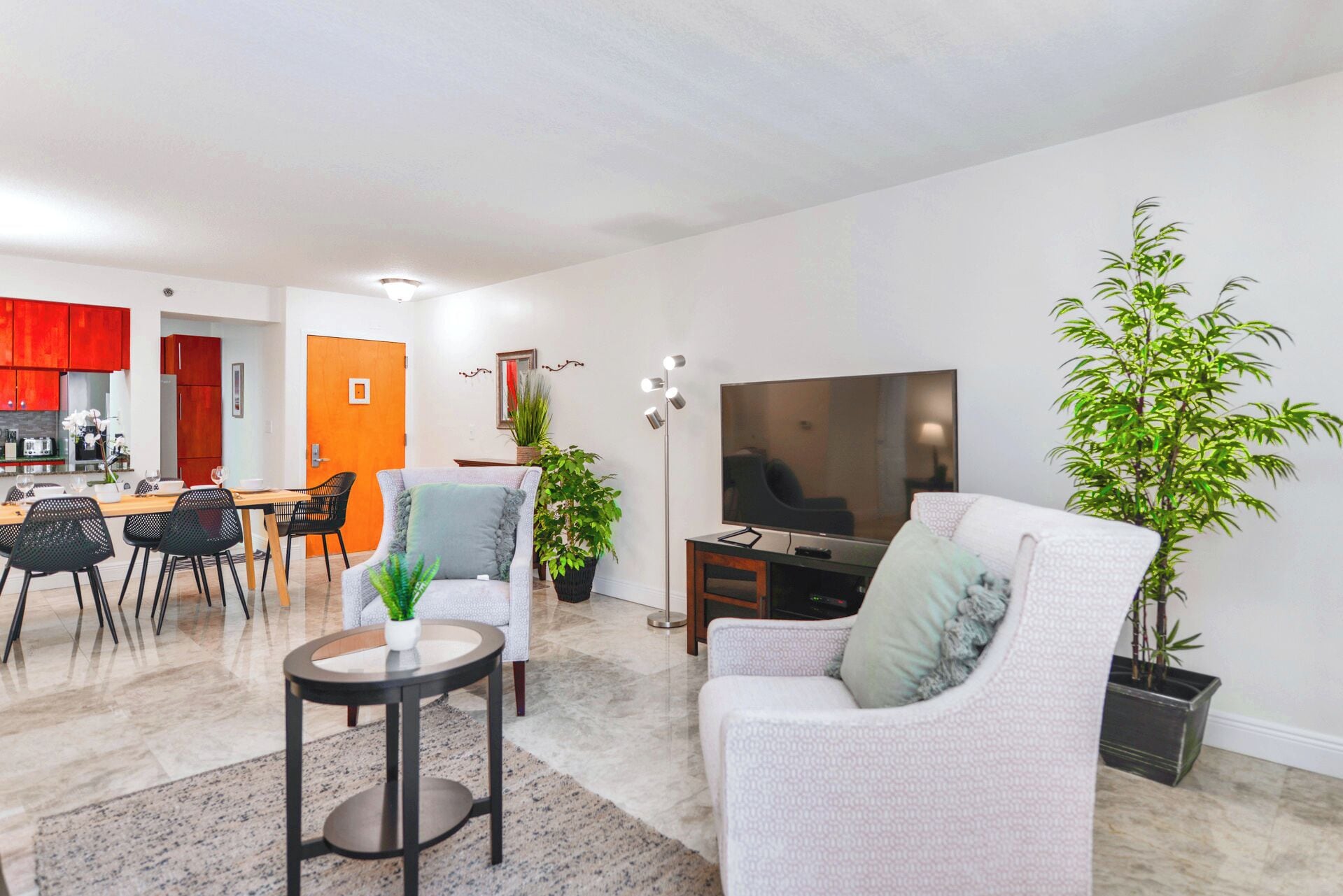 A modern living room with a flat-screen TV, a sofa, a dining area with a table and chairs, and a kitchen in the background.