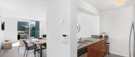 A modern kitchen with a dishwasher, sink, and wooden cabinets. There is a dining area with a table and chairs, and a view of the city through large windows.