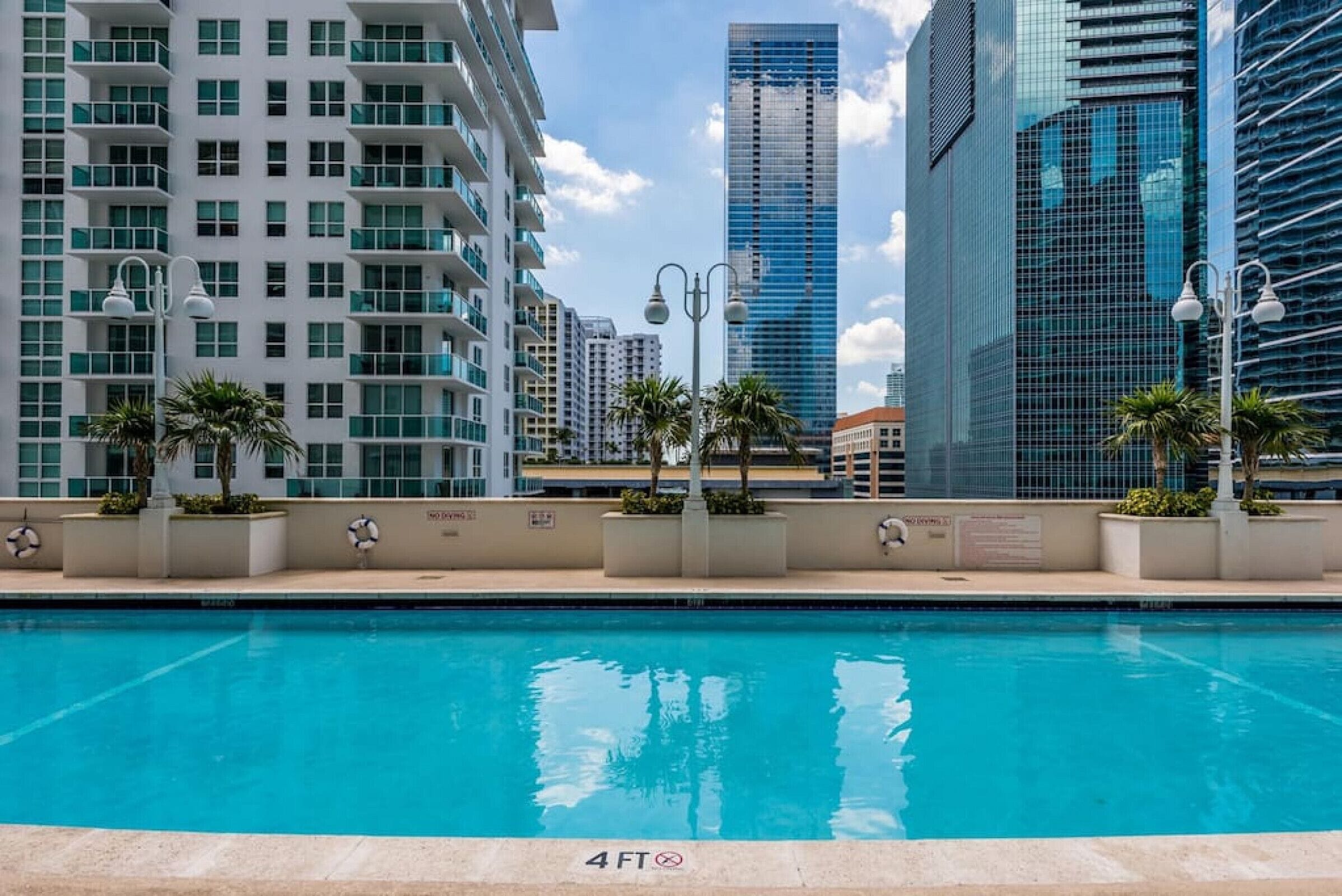 Piscina con límite de profundidad de 1.2 metros, rodeada de edificios modernos y palmeras.