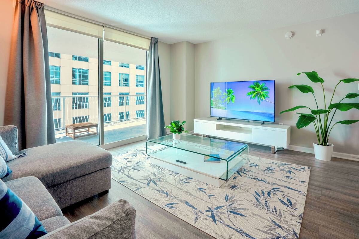 A modern living room with a sofa, a glass coffee table, a flat-screen TV, a potted plant, and a view of a building through large windows.
