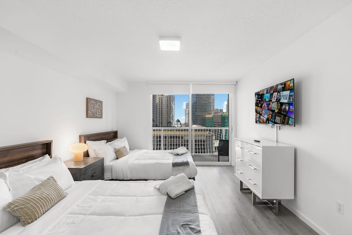A modern bedroom with two beds, a TV, and a balcony with a city view.