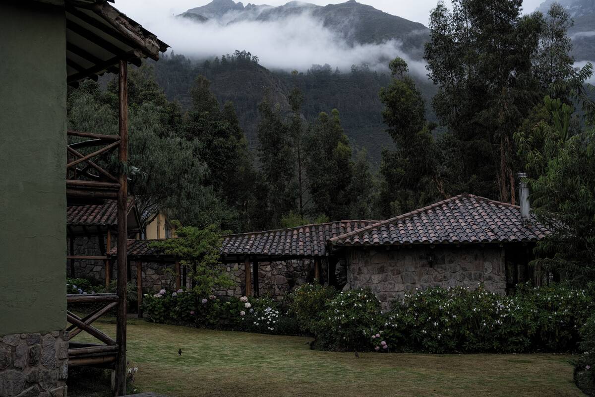 A rustic stone building with a tiled roof, surrounded by lush greenery and a mountainous backdrop.