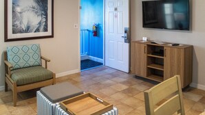 A room with a framed picture, a television, a wooden cabinet, a chair, and a door leading to a blue hallway. - Barefoot'n Resort Kissimmee Orlando (Kissimmee)