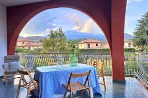 A balcony with a table set for two, a chair, and a view of houses and mountains in the background. - The Little Vineyard (Trecastagni)