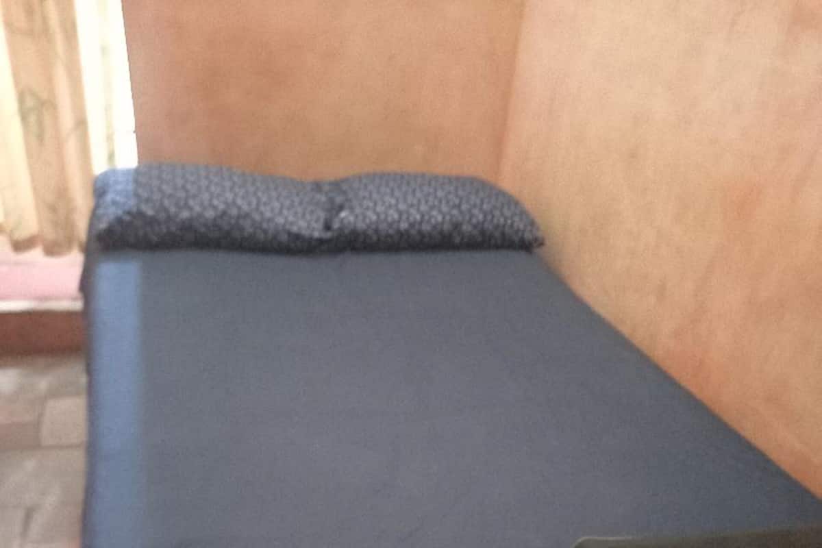 Ein kleines Zimmer mit einem Einzelbett, einer dunklen Bettdecke und passendem Kopfkissen. Der Raum hat einen Fliesenboden und eine schlichte Wand.