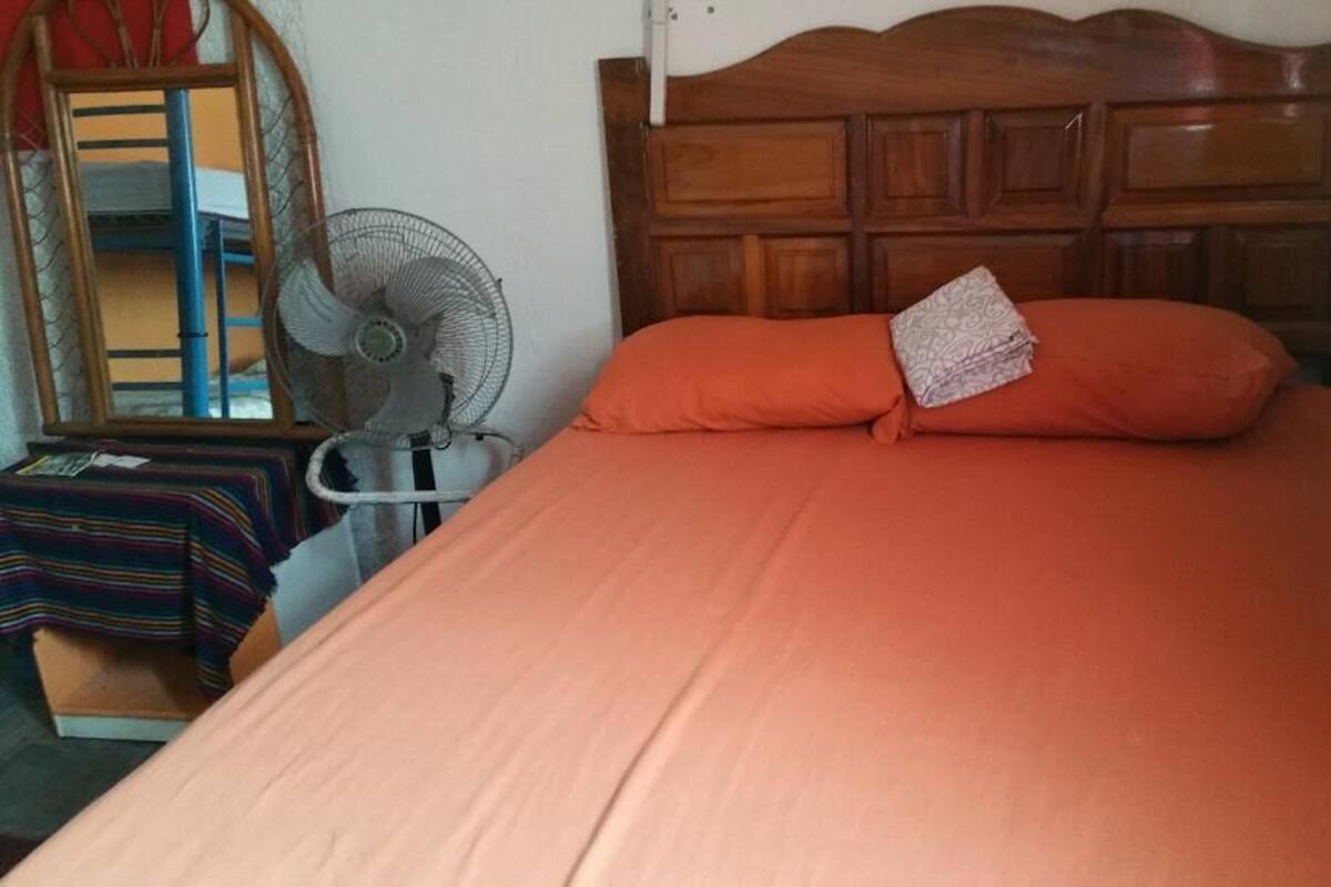 Ein Schlafzimmer mit einem hölzernen Kopfteil, einer orangefarbenen Bettdecke und einem Ventilator auf einem kleinen Tisch.