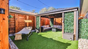 Een houten terras met een tafel en stoelen, een bankje, en een overdekte zithoek met een groene wand.