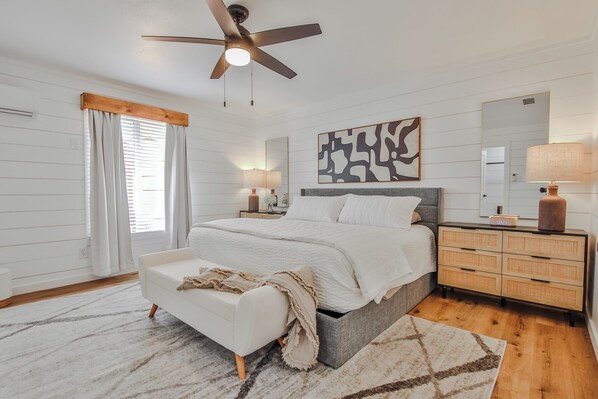 A bedroom with a bed, bedside tables, a ceiling fan, a large mirror, and a window with curtains.