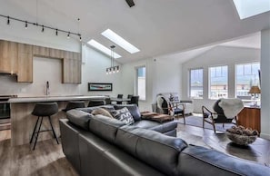 A modern kitchen with a bar area, a large leather sofa, and a dining table in a spacious living area with large windows.