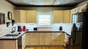 Une cuisine avec un plancher en bois, des armoires de couleur beige, un réfrigérateur en acier inoxydable et une fenêtre avec des stores.