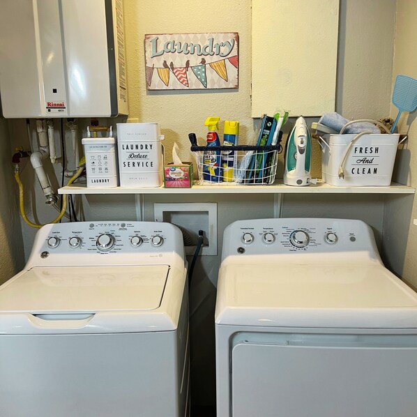 Une buanderie équipée de deux lave-linge et deux sèche-linge, avec un panneau indiquant qu’il s’agit d’une laverie en libre-service, et divers produits de nettoyage rangés sur des étagères.