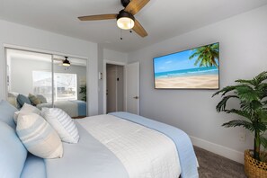 A bedroom with a bed, a ceiling fan, a beach-themed wall art, and a potted plant.