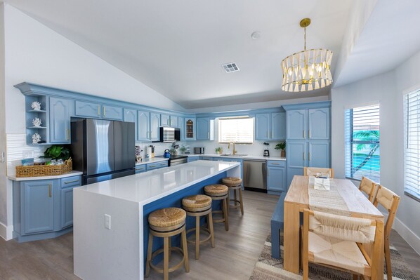 A modern kitchen with blue cabinets, a large island with stools, stainless steel appliances, and a dining area with wooden chairs and a table.