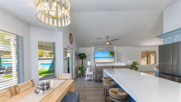 A modern kitchen with a large island, a dining area with a bench, and a living room with a sofa and a TV displaying a beach scene. - Seaside Cottage in walking distance of downtown Gilbert with charming pool. (Gilbert)