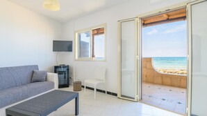 Een moderne woonkamer met een grijze bank, een kleine tafel, een televisie en een schuifdeur met uitzicht op het strand.