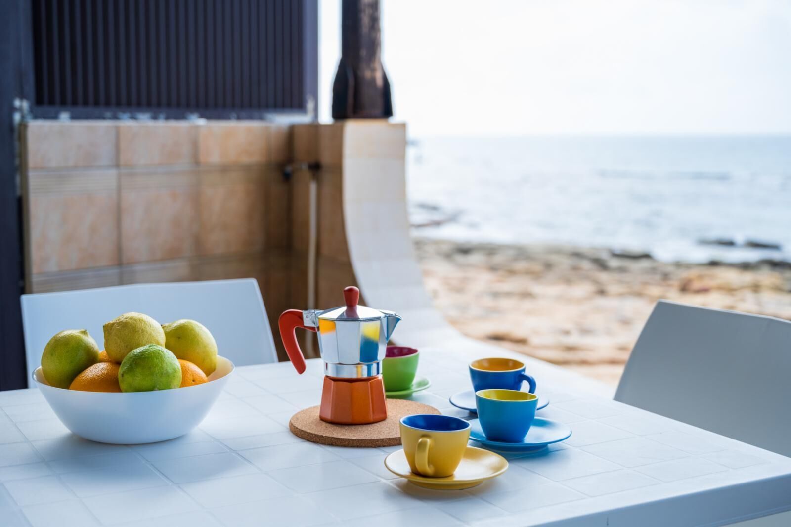 A table with a coffee maker, cups, and a bowl of fruit on a terrace overlooking the sea.