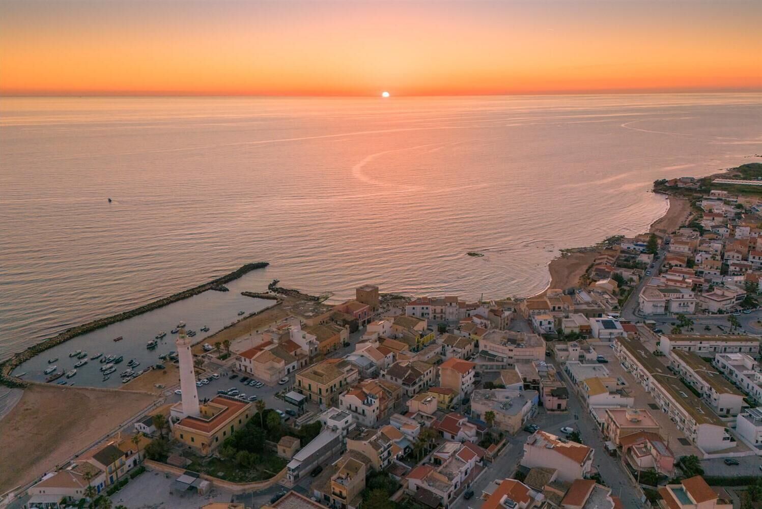 Une ville côtière au coucher du soleil, avec un phare imposant, une marina remplie de bateaux et un mélange de bâtiments résidentiels.