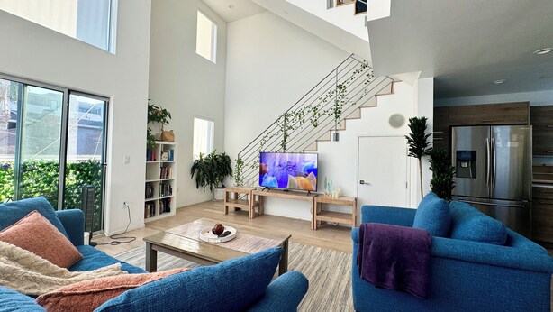 A spacious living room with a large TV, blue sofas, a staircase, and a kitchen area. - Skydeck MidWilshire Modern Townhome with Garage, Rooftop Patio, and Loft Office (Los Angeles)
