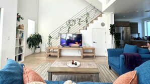 A spacious living room with a staircase, a television, and a kitchen area. - Skydeck MidWilshire Modern Townhome with Garage, Rooftop Patio, and Loft Office (Los Angeles)