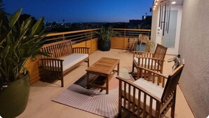 A balcony with wooden furniture, a potted plant, and a view of the city at dusk. - Skydeck MidWilshire Modern Townhome with Garage, Rooftop Patio, and Loft Office (Los Angeles)